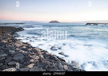 Alba sulla spiaggia, rocce laviche e spiaggia sabbiosa in lunga esposizione. Posto romantico con molo al mattino. Playa de la Barreta, Parco Nazionale di Corralejo, Isole Canarie, Spagna Foto Stock