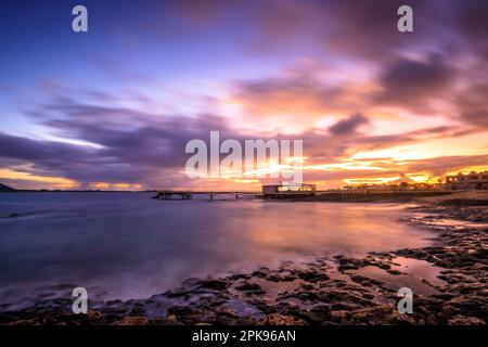 Alba sulla spiaggia, rocce laviche e spiaggia sabbiosa in lunga esposizione. Posto romantico con molo al mattino. Playa de la Barreta, Parco Nazionale di Corralejo, Isole Canarie, Spagna Foto Stock