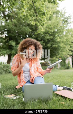 donna allegra con capelli ricci che tiene il giornale e il caffè per andare mentre si siede sull'erba e guardando il portatile, immagine di scorta Foto Stock