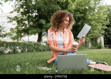 donna allegra con capelli ricci leggere giornale e tenere il caffè per andare mentre si siede sull'erba vicino al computer portatile, immagine stock Foto Stock