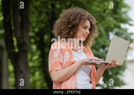 allegra donna freelance sorridente mentre usa il notebook e lavora in remoto in parco, immagine stock Foto Stock