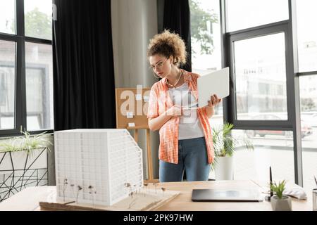 design architettonico riccio che tiene il computer portatile mentre si guarda il modello di casa in ufficio, immagine di scorta Foto Stock