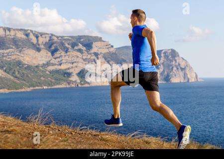 sentiero montano da runner maschio lungo la riva dell'oceano, abbigliamento sportivo, camicia blu e pantaloncini neri Foto Stock