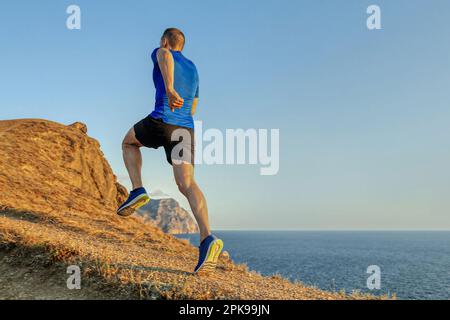 torna maschio runner correre in salita sentiero di montagna su sfondo cielo blu, jogging estivo lungo la costa al tramonto Foto Stock