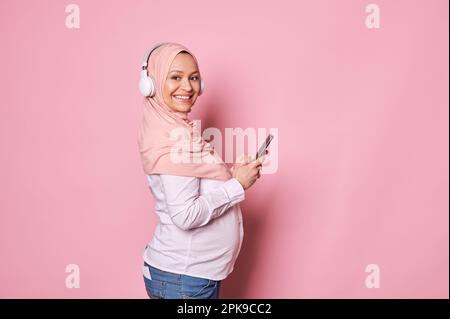 Sorridente donna musulmana incinta felice in hijab rosa e cuffie wireless, controllando la nuova applicazione mobile sullo smartphone Foto Stock