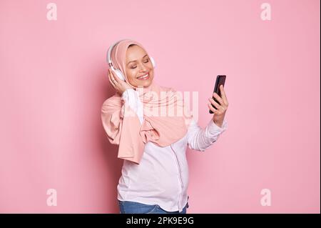 Donna musulmana incinta in hijab rosa e cuffie, gode di una musica rilassante, posando con smartphone su sfondo rosa Foto Stock