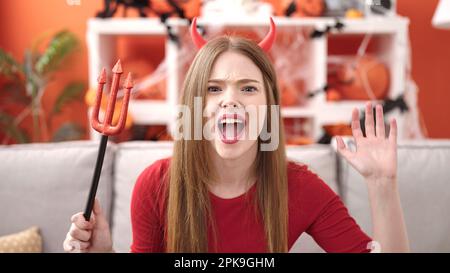 Giovane donna bionda che indossa un costume da diavolo facendo un gesto di paura a casa Foto Stock