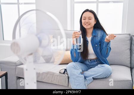 Giovane donna asiatica bere un bicchiere d'acqua godendo l'aria dal ventilatore urlando fiero, celebrando la vittoria e il successo molto eccitato con il braccio sollevato Foto Stock