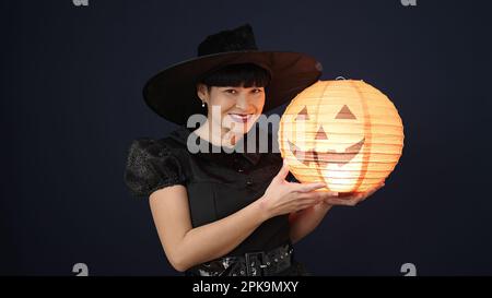 Giovane donna cinese che indossa il costume della strega che tiene la lampada della zucca di Halloween sopra lo sfondo nero isolato Foto Stock