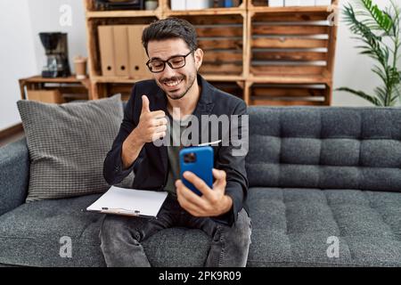 Giovane psicologo ispanico uomo che fa la terapia in videochiamata con smartphone sorridente felice e positivo, pollice su facendo eccellente e segno di approvazione Foto Stock