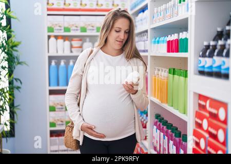 Giovane donna incinta cliente che tocca la pancia tenendo pillole scaffalatura bottiglia in farmacia Foto Stock