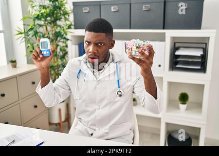 Dottore afro-americano che usa il glucometro in faccia d'urto, che guarda scettico e sarcastico, sorpreso dalla bocca aperta Foto Stock