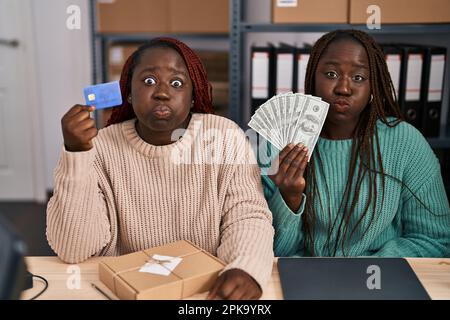 Due donne africane che lavorano a piccolo commercio elettronico che tiene la carta di credito e le banconote puffing guance con faccia divertente. bocca gonfiata con aria, presa Foto Stock