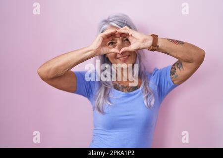 Donna di mezza età con tatuaggi in piedi su sfondo rosa facendo forma di cuore con mano e dita sorridenti guardando attraverso il segno Foto Stock