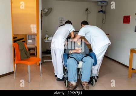 Due assistenti infermieri mettono un paziente disabili a letto nella sua stanza presso la casa di cura. Foto Stock