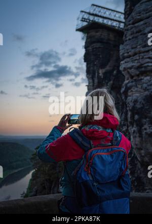 Montagne di arenaria dell'Elba Sassonia Svizzera: Impressioni. Donna bionda con zaino fotografare il fiume Elba con telefono cellulare, vista posteriore. Foto Stock