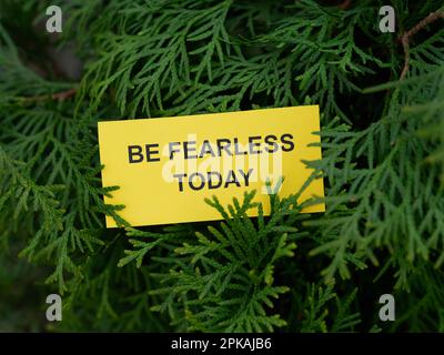 Una nota di carta gialla con la frase essere senza orecchio oggi su di essa appesa su un albero di arborvitae. Primo piano. Foto Stock