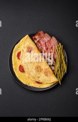 Deliziosa omelette fresca con pomodori ciliegini, pancetta, asparagi e spezie su fondo di cemento scuro Foto Stock