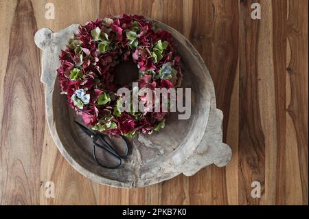 Corona fiorita legata da fiori di idrangea rosso scuro e verde, sistemata in vecchia ciotola di legno con forbici da fiore, decorazione con materiali naturali Foto Stock