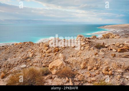 Riva del mare morto sul lato del Giordano, spiaggia di sabbia asciutta e rocce, il sole splende su una bellissima superficie di acqua azzurra Foto Stock