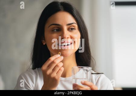 Multirazziale asiatica sorridente giovane donna che prende una capsula di olio di pesce Omega-3 a casa con bei denti bianchi, sano stile di vita cibo dietetico integratori Foto Stock