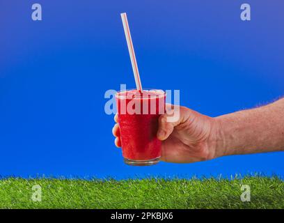 Frullato di frutta con una paglia in esso, essendo tenuto fuori sopra un certo erba con uno sfondo blu del cielo. Foto Stock