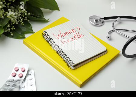 Notebook con testo Women's Health, pillole e stetoscopio su tavolo bianco Foto Stock