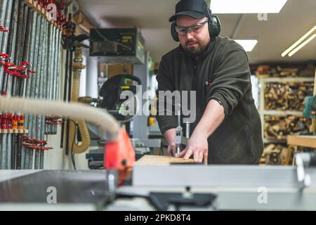 Interni di officina di lavorazione del legno. Artigiano focalizzato in occhiali protettivi e cuffie utilizzando la sega da tavolo. . Foto di alta qualità Foto Stock