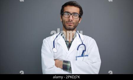 Primo piano di un giovane medico maschile in un cappotto bianco e bicchieri tondi isolati su uno sfondo grigio Foto Stock