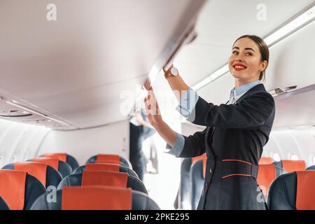 Giovane stewardess che è in abiti neri formali è in piedi all'interno dell'aereo Foto Stock