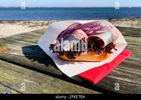 Un panino di pesce ripieno di giovani aringhe salate si trova su un tavolo in spiaggia. Foto Stock