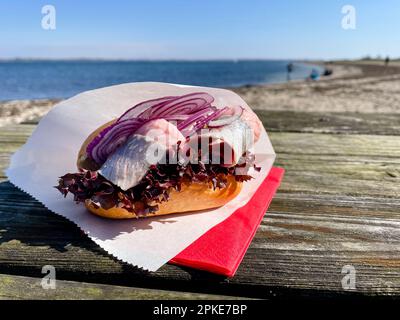 Un panino di pesce ripieno di giovani aringhe salate si trova su un tavolo in spiaggia. Foto Stock