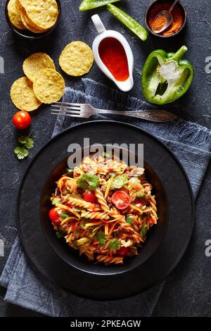 Insalata di pasta Nacho con carne macinata, pomodori, peperoni verdi, lattuga iceberg e condimento taco in ciotola nera su tavolo grigio in cemento, vista verticale fr Foto Stock