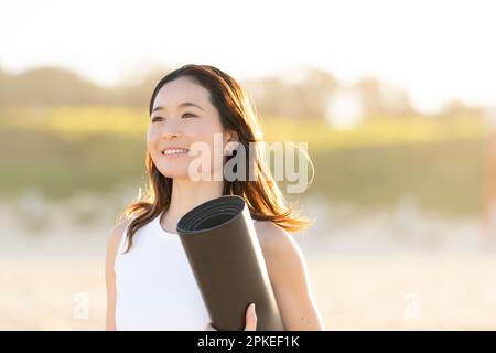Donna che cammina sulla spiaggia con un tappetino yoga Foto Stock