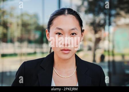 Primo piano ritratto di una giovane donna asiatica d'affari o dirigente che guarda seriamente la fotocamera indossando una tuta in piedi all'esterno dell'area di lavoro. Vista frontale della giovane avvocato con atteggiamento penitente. . Foto di alta qualità Foto Stock