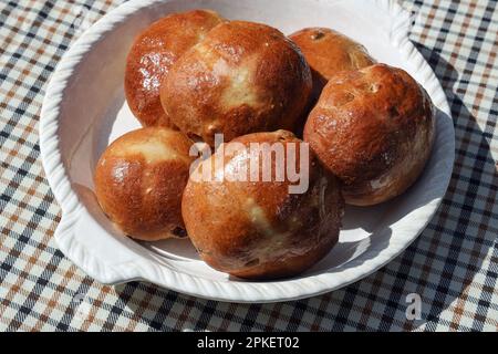 Un semplice piatto di grandi panini a croce caldi fatti a mano con uno smalto di zucchero su una tovaglia di gingham marrone e nero. Foto Stock