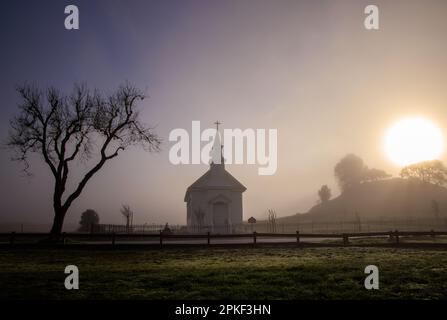 Sole luminoso e nebbia scura sopra la piccola chiesa e l'albero nella campagna all'alba Foto Stock