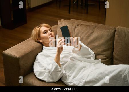 Bella bionda di mezza età si trova su un divano in un accappatoio Foto Stock