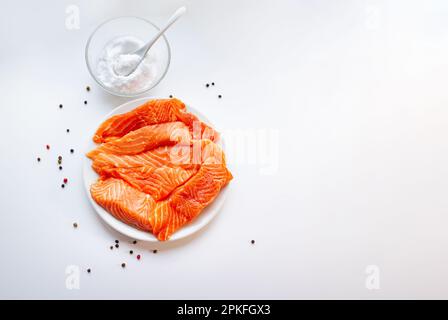 Filetto di salmone fresco crudo su un piatto bianco, una piccola ciotola di sale e pepe nero su fondo bianco. Cibo sano. Foto Stock