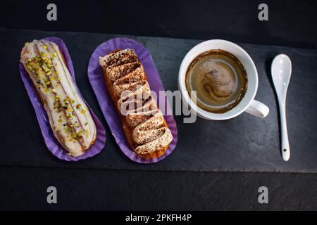 Tazza di caffè e deliziose torte eclair - tiramisù e pistacchio aromatizzati con panna, glassa e noci tritate, su fondo nero. Foto Stock