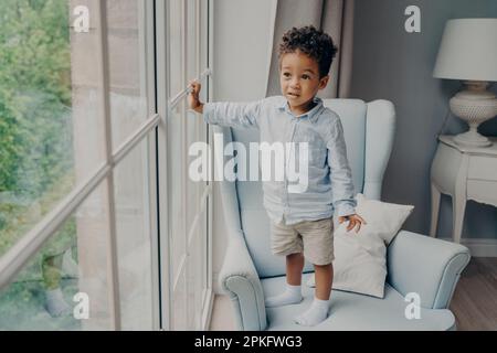 Lunghezza completa di piccolo afro curioso americano ragazzo dolce toddler in stile di indossare guardando attraverso la finestra e in attesa di mamma, mentre trascorrere il tempo libero a. Foto Stock