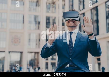 Un giovane uomo d'ufficio felice vestito formalmente seduto all'aperto e utilizzando visore VR o occhiali 3D, interagendo con l'interfaccia digitale e con exp Foto Stock