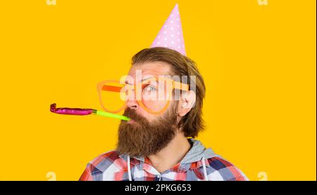 Feste e celebrazioni. Uomo bearded in cappello di compleanno e divertenti occhiali che soffiano il fischio di festa. Buon compleanno. Bell'uomo in cappello conico e grande Foto Stock