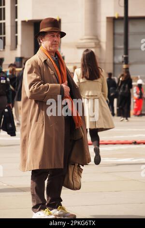 Un uomo più anziano 60+ in piedi con cappotto lungo, borsa grande, sciarpa arancione, cappello marrone in piedi a Trafalgar Square, Londra guardando e ascoltando un busker Foto Stock