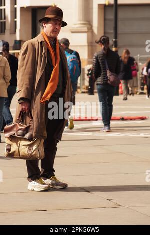 Un uomo più anziano 60+ in piedi con cappotto lungo, borsa grande, sciarpa arancione, cappello marrone in piedi a Trafalgar Square, Londra guardando e ascoltando un busker Foto Stock