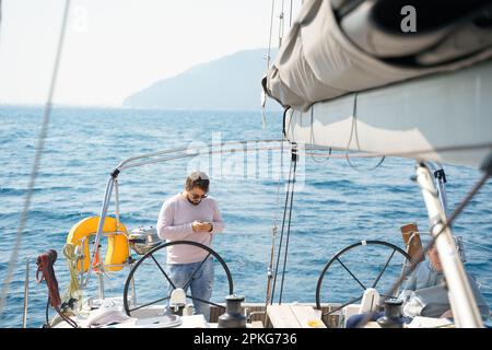 Uomo d'affari, proprietario con telefono cellulare al volante in viaggio vela su sport mare yacht di lusso. Crociera estiva in barca. Vela sull'oceano shi Foto Stock