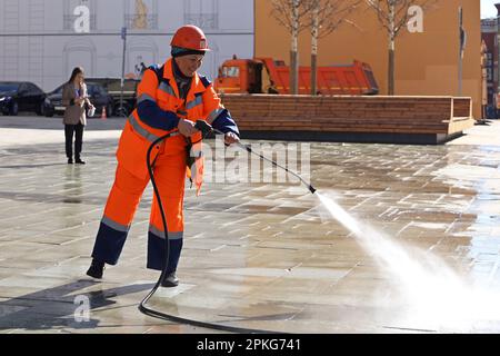 Donna asiatica felice in uniforme di lavoro che innaffia il marciapiede con un tubo flessibile. Pulizia della strada e disinfezione nella città di primavera Foto Stock