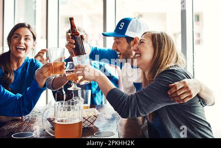 Alzando i loro occhiali ad un gioco impressionante. un gruppo di amici brinda con le birre mentre guarda una partita sportiva in un bar. Foto Stock