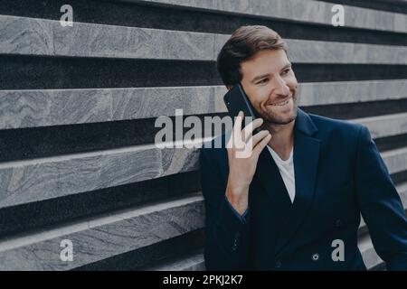 Giovane lavoratore in ufficio attraente in blazer elegante parlare sul telefono cellulare durante la pausa di lavoro, sorridendo sinceramente mentre la famiglia piacevole parlare su smartp Foto Stock