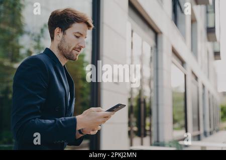 Bell'uomo concentrato con barba che indossa abiti scuri che chiacchierano sullo smartphone, navigando sul Web sul telefono mentre si è in piedi davanti all'edificio con un sacco di vento Foto Stock